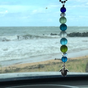 May include: A car charm with a chain of dark beads and a series of colourful beads in blue, green, and white. A small silver turtle charm hangs at the bottom. The background shows a beach and ocean.