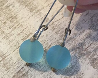 Puede incluir: Un par de pendientes con cuentas redondas de color azul claro suspendidas de largos alambres plateados. Los pendientes se muestran sobre una superficie de madera clara y texturizada.