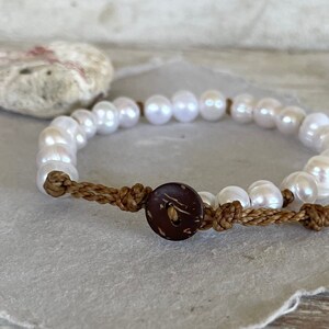May include: A white pearl bracelet with a brown coconut button closure. The bracelet is made with a brown cord and is tied with a knot.