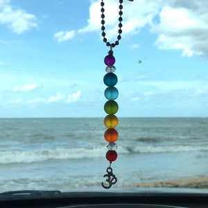 May include: A car charm featuring a string of colorful, frosted glass beads in a rainbow spectrum, with a silver-toned Om symbol pendant. The beads are purple, clear, blue, green, yellow, orange, and red. The charm hangs in front of a seascape.