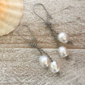 May include: A pair of handmade earrings featuring large, lustrous white pearls suspended from gray woven cords. The earrings have silver-colored ear wires and are displayed on a light-colored wooden surface, with a seashell in the background.