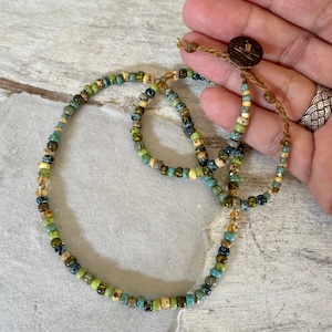 May include: A beaded necklace featuring a mix of small, colorful beads in shades of green, blue, and cream. The necklace is strung on a brown cord and has a wooden button closure. The beads are arranged in a repeating pattern.