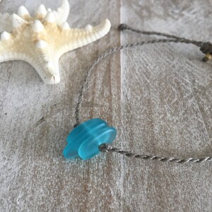 Puede incluir: Un collar con un cordón gris y una cuenta de vidrio azul. La cuenta está hecha de piezas redondas de vidrio apiladas.