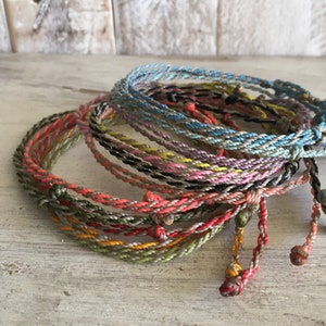 May include: A stack of colorful braided bracelets with knotted ends. The bracelets are made of thin, twisted cords in various shades of blue, green, pink, yellow, and orange.