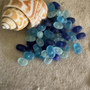 May include: A seashell with brown and white stripes beside a collection of blue glass beads. The beads range in shades from light to dark blue. The items are arranged on a textured, light-coloured surface.