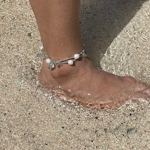 May include: A silver anklet with white pearls and clear glass beads. The anklet is worn on a person's ankle, and the foot is submerged in shallow water.