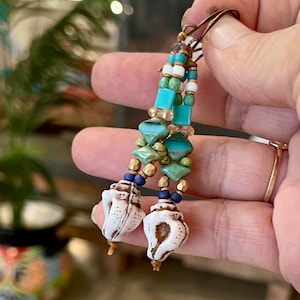 Könnte beinhalten: Ein Paar lange, geperlte Ohrringe mit einem Muschel-Anhänger am Ende. Die Perlen sind in Blautönen, Grün- und Goldtönen.