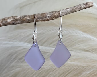 Puede incluir: Un par de pendientes en forma de diamante de color lavanda con alambres plateados. Los pendientes cuelgan de una pequeña rama marrón claro sobre un fondo suave y borroso.