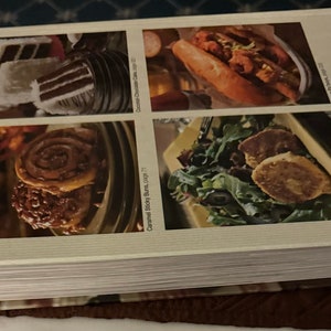 May include: A cookbook with a cream-colored cover featuring four food images: a slice of cake, a sandwich, cinnamon rolls, and a salad. The book's title is not visible, but the publisher's name, "Oxmoor House," is printed on the spine.