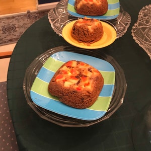 May include: Three small, round cakes with fruit toppings are presented on colorful plates. The cakes sit on plates with blue and green stripes, and a yellow plate. The cakes are on a dark green tablecloth.