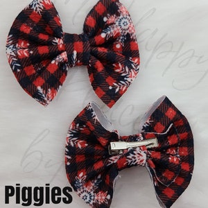May include: Two red and black plaid fabric hair bows with white snowflake accents. One bow is positioned above the other. The bottom bow has a silver metal clip. The word "Piggies" is printed in black at the bottom.