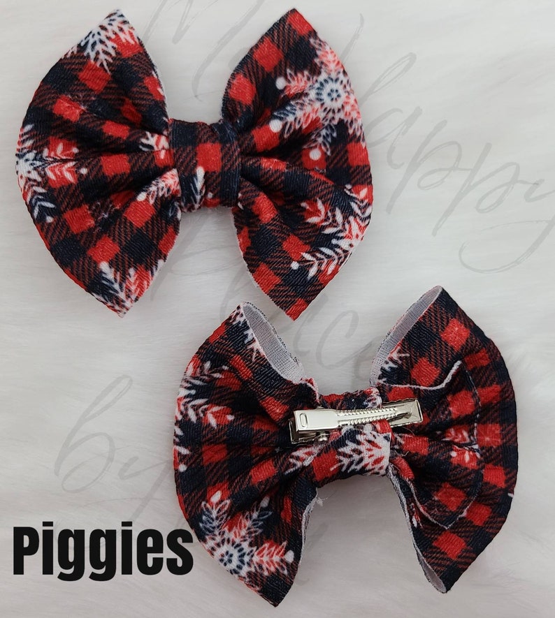 May include: Two red and black plaid fabric hair bows with white snowflake accents. One bow is positioned above the other. The bottom bow has a silver metal clip. The word "Piggies" is printed in black at the bottom.