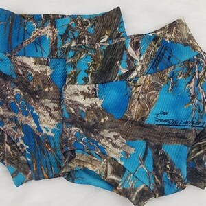 May include: Two pairs of blue camouflage print shorts. The shorts feature a high waistband and a pattern of blue, brown, and gray resembling a forest. The fabric has a ribbed texture. The shorts have a signature on the side.