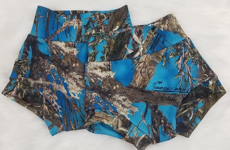 May include: Two pairs of blue camouflage print shorts. The shorts feature a high waistband and a pattern of blue, brown, and gray resembling a forest. The fabric has a ribbed texture. The shorts have a signature on the side.