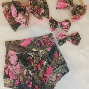 May include: A set of baby clothing and accessories in a pink and brown camouflage pattern. The set includes a pair of shorts, a headband, and several bow hair clips. The items are arranged on a white surface.