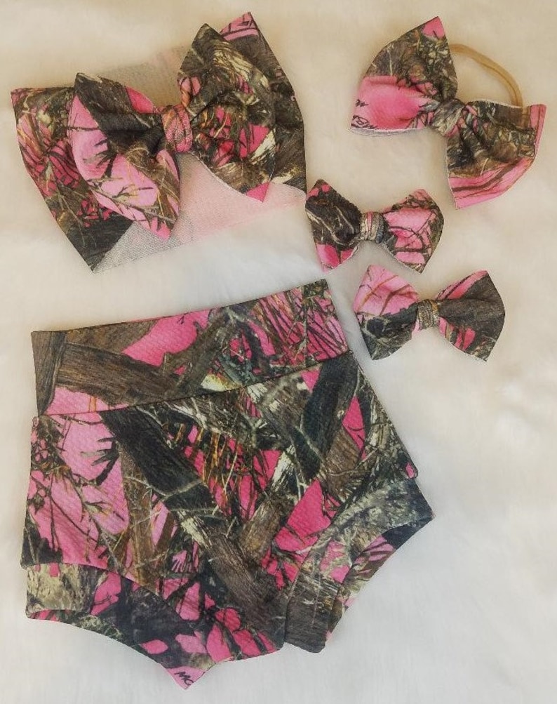 May include: A set of baby clothing and accessories in a pink and brown camouflage pattern. The set includes a pair of shorts, a headband, and several bow hair clips. The items are arranged on a white surface.