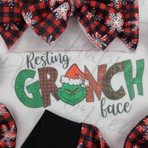 May include: A white onesie with the text "Resting Grinch face" and a Grinch graphic, surrounded by red and black plaid bows with white snowflake accents. A black band is also visible.
