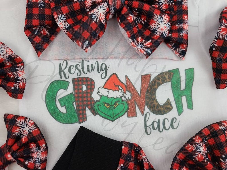 May include: A white onesie with the text "Resting Grinch face" and a Grinch graphic, surrounded by red and black plaid bows with white snowflake accents. A black band is also visible.