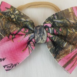 May include: A pink and camouflage bow with a beige elastic band. The bow features a vibrant pink color combined with a camouflage pattern in shades of brown, green, and black. The bow is tied in the center.