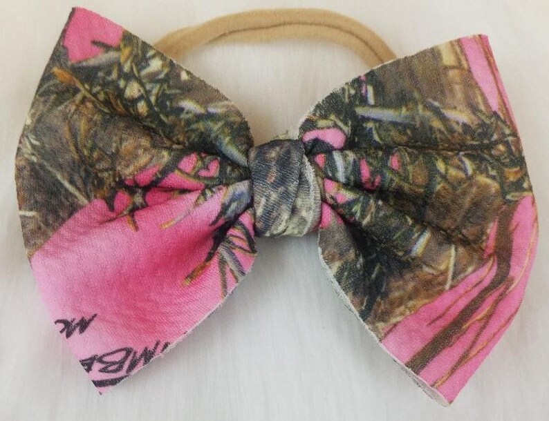 May include: A pink and camouflage bow with a beige elastic band. The bow features a vibrant pink color combined with a camouflage pattern in shades of brown, green, and black. The bow is tied in the center.