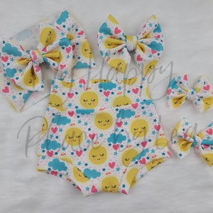 May include: A set of three white, yellow, pink, and blue fabric hair bows with a sun and cloud pattern. The bows are made of a soft, textured fabric and are perfect for adding a touch of whimsy to any outfit.