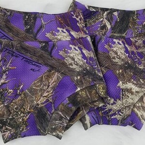 May include: Two pairs of purple and brown camouflage print shorts. The shorts have a ribbed texture and a wide waistband. The camouflage pattern features shades of purple, brown, and white, creating a unique and eye-catching design.