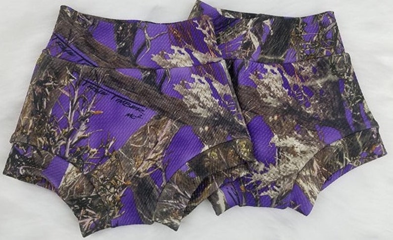 May include: Two pairs of purple and brown camouflage print shorts. The shorts have a ribbed texture and a wide waistband. The camouflage pattern features shades of purple, brown, and white, creating a unique and eye-catching design.