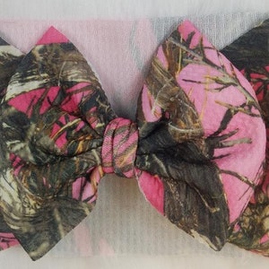 May include: A pink and camouflage fabric bow headband. The bow features a pink and brown camouflage pattern. The headband is made of a soft, stretchy material. The bow is tied in the center and is a large size.