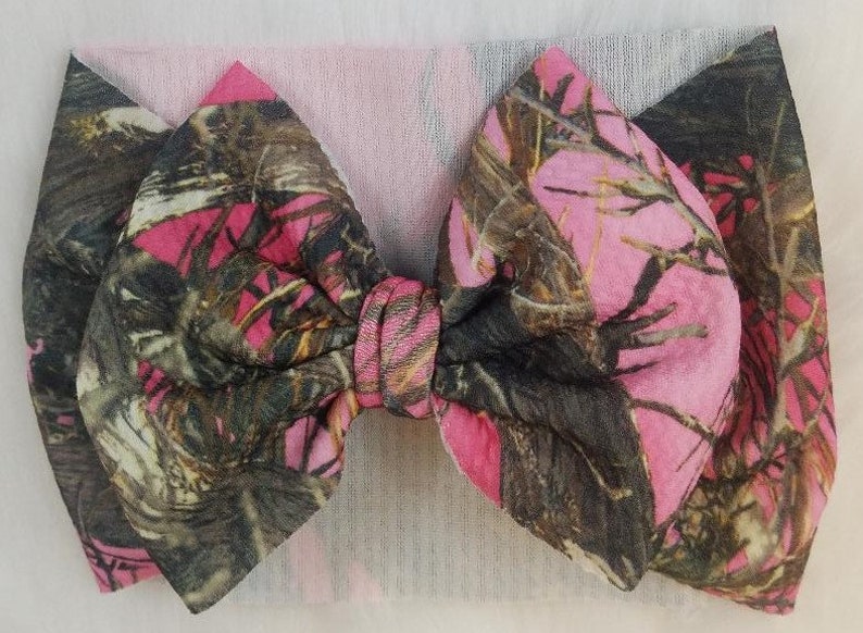 May include: A pink and camouflage fabric bow headband. The bow features a pink and brown camouflage pattern. The headband is made of a soft, stretchy material. The bow is tied in the center and is a large size.