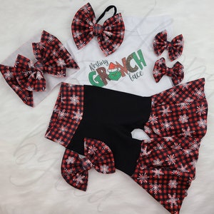 May include: A festive baby outfit set featuring a white onesie with a Grinch graphic and the text "Resting Grinch Face." The set includes black shorts with red and black plaid snowflake trim, and several matching plaid bows.