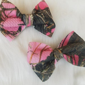 May include: Two fabric hair bows with a pink and camouflage pattern. The bows are made of a soft material and have a classic bow shape. The bows are likely for children or babies.