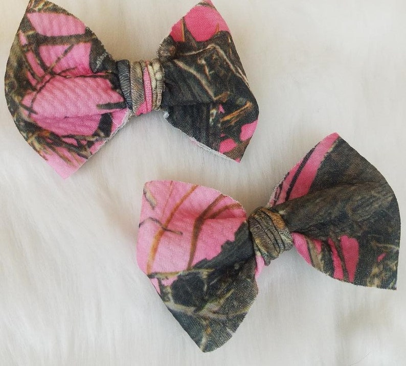 May include: Two fabric hair bows with a pink and camouflage pattern. The bows are made of a soft material and have a classic bow shape. The bows are likely for children or babies.