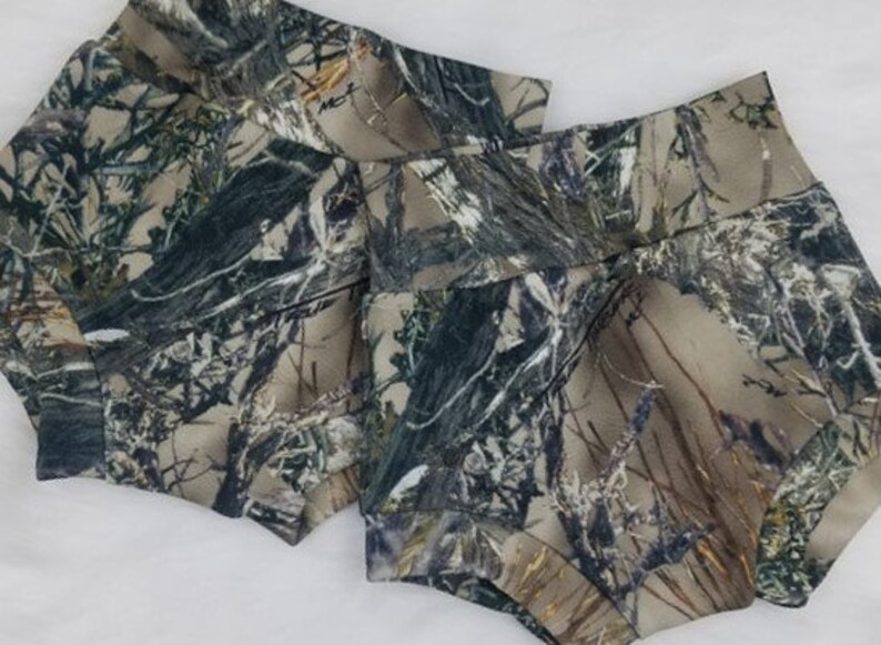May include: Two pairs of camouflage print shorts. The shorts feature a high waist and are made of a fabric with a pattern of green, brown, and tan hues, resembling a forest environment. The shorts are designed for comfort and style.