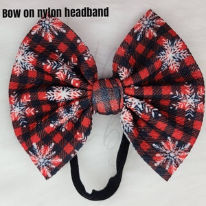 May include: A large, decorative bow on a black nylon headband. The bow features a red and black plaid pattern with white snowflake accents. The text "Bow on nylon headband" is visible at the top of the image.
