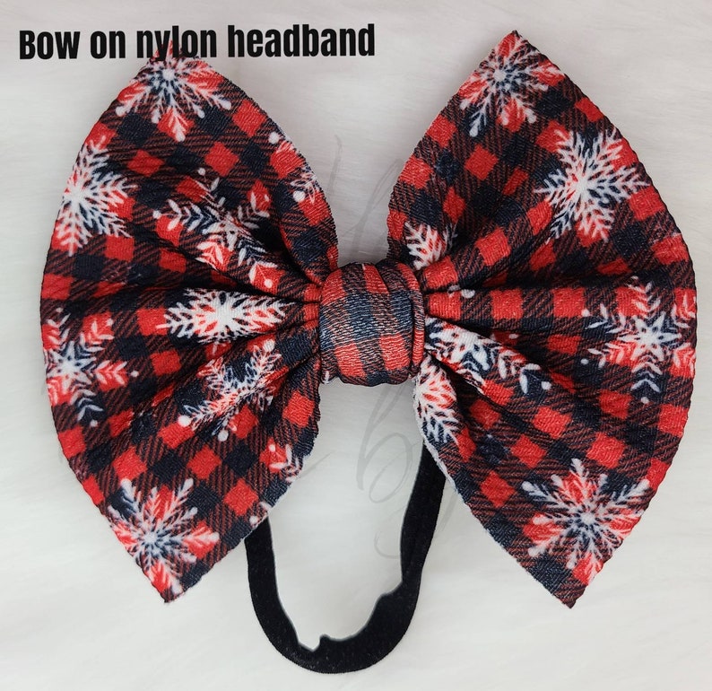 May include: A large, decorative bow on a black nylon headband. The bow features a red and black plaid pattern with white snowflake accents. The text "Bow on nylon headband" is visible at the top of the image.