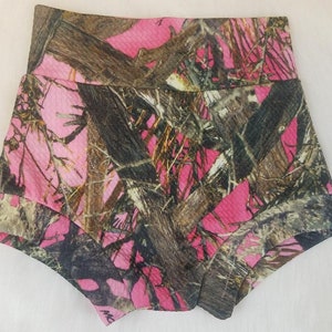 May include: Pink camouflage baby shorts with a high waistband and layered design. The shorts feature a pattern of pink, brown, and black, resembling a woodland camouflage print. The fabric appears to have a textured surface.