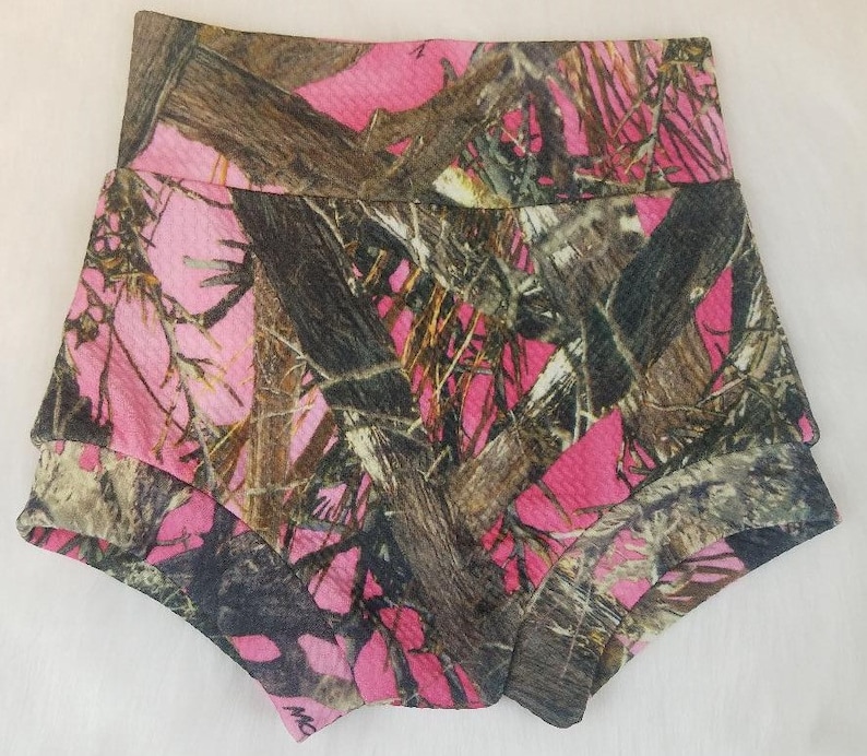 May include: Pink camouflage baby shorts with a high waistband and layered design. The shorts feature a pattern of pink, brown, and black, resembling a woodland camouflage print. The fabric appears to have a textured surface.