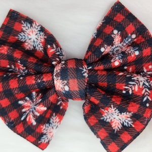 May include: A large, fabric hair bow featuring a red and black buffalo plaid pattern. White snowflake designs are scattered across the bow. The bow is tied in the center with a matching plaid knot, creating a festive accessory.
