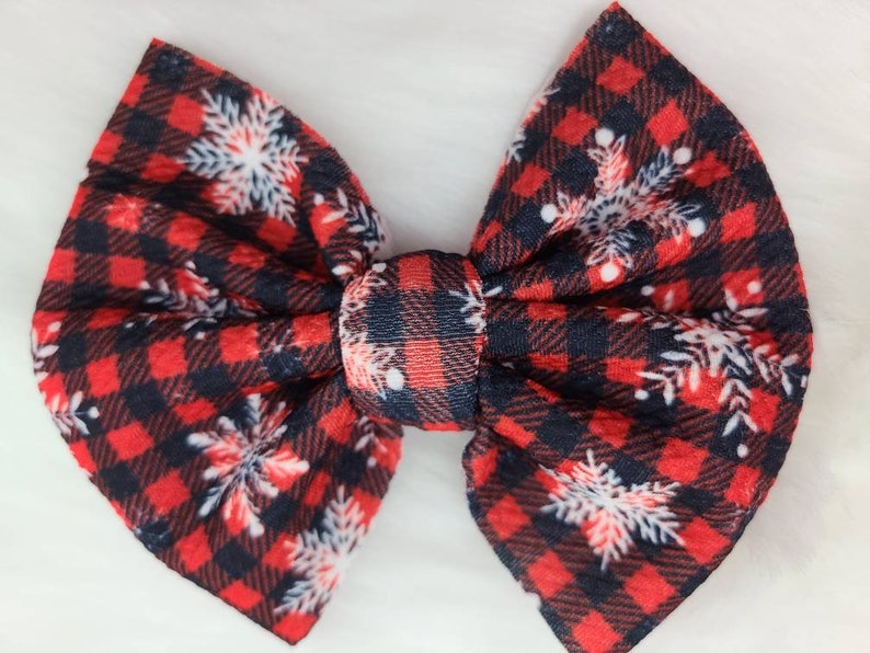 May include: A large, fabric hair bow featuring a red and black buffalo plaid pattern. White snowflake designs are scattered across the bow. The bow is tied in the center with a matching plaid knot, creating a festive accessory.