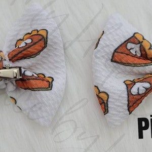 May include: Two white hair bows with a clip on the back. The bows are made of a white fabric with a pattern of orange and brown pie slices. The text "Piggies" is printed on the bottom right of the image.
