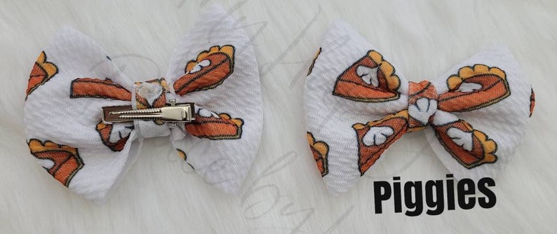 May include: Two white hair bows with a clip on the back. The bows are made of a white fabric with a pattern of orange and brown pie slices. The text "Piggies" is printed on the bottom right of the image.