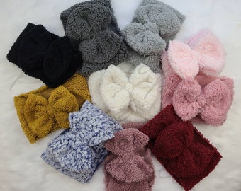 May include: Assortment of fuzzy headbands in various colors, including pink, gray, brown, blue, and red. Each headband features a large bow.