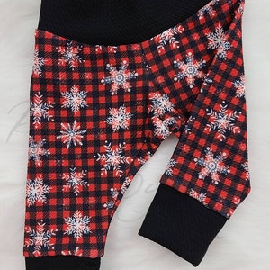 May include: Baby leggings with a red and black plaid pattern, adorned with white and blue snowflakes. The leggings have a black waistband and cuffs. The fabric appears soft and comfortable, suitable for winter wear.