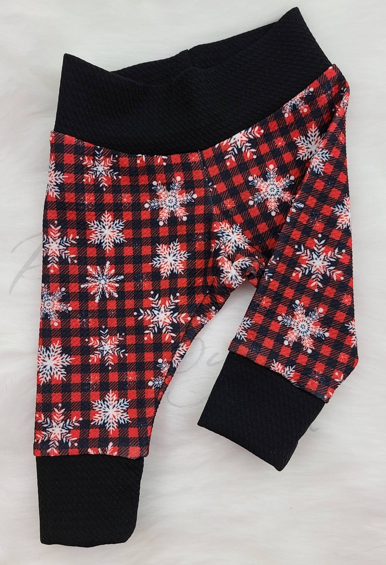 May include: Baby leggings with a red and black plaid pattern, adorned with white and blue snowflakes. The leggings have a black waistband and cuffs. The fabric appears soft and comfortable, suitable for winter wear.