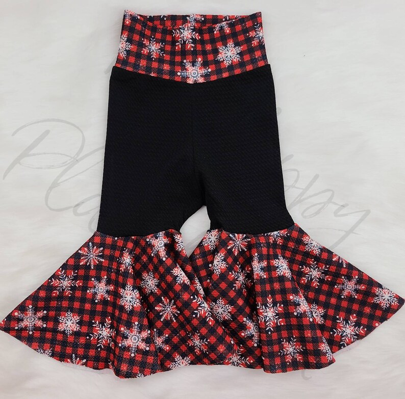May include: Black bell-bottom pants with a red and black plaid snowflake pattern on the waistband and flared bottoms. The pants have a textured fabric and a high waist design, perfect for holiday or winter wear.