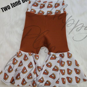 May include: Brown and white bell bottom trousers with a pumpkin pie print. The trousers have a brown waffle knit texture and a white pumpkin pie print on the bottom.