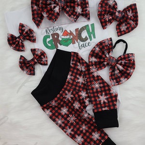 May include: A festive baby outfit set featuring a white onesie with a Grinch graphic and the text "Resting Grinch Face." The set includes red and black plaid snowflake print leggings, and several matching hair bows.