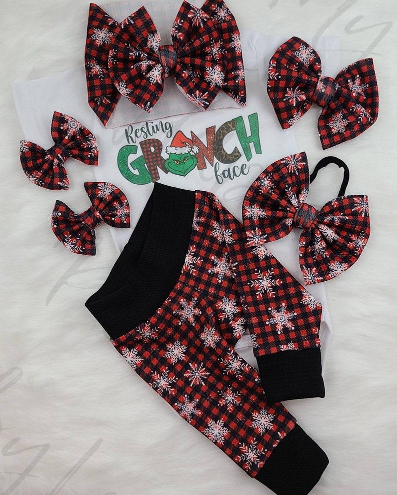 May include: A festive baby outfit set featuring a white onesie with a Grinch graphic and the text "Resting Grinch Face." The set includes red and black plaid snowflake print leggings, and several matching hair bows.