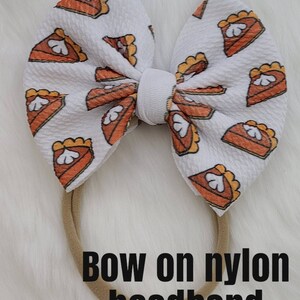 May include: White bow with a pumpkin pie print on a brown nylon headband. "Bow on nylon headband" is written below the bow.