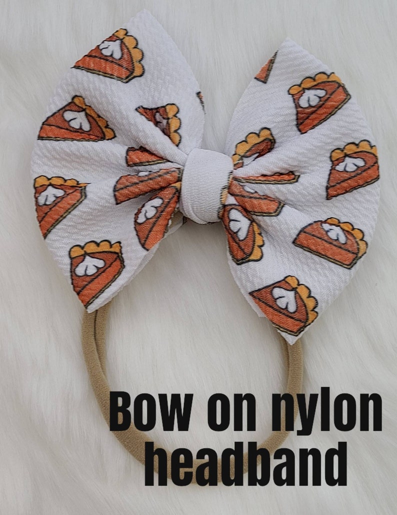 May include: White bow with a pumpkin pie print on a brown nylon headband. "Bow on nylon headband" is written below the bow.